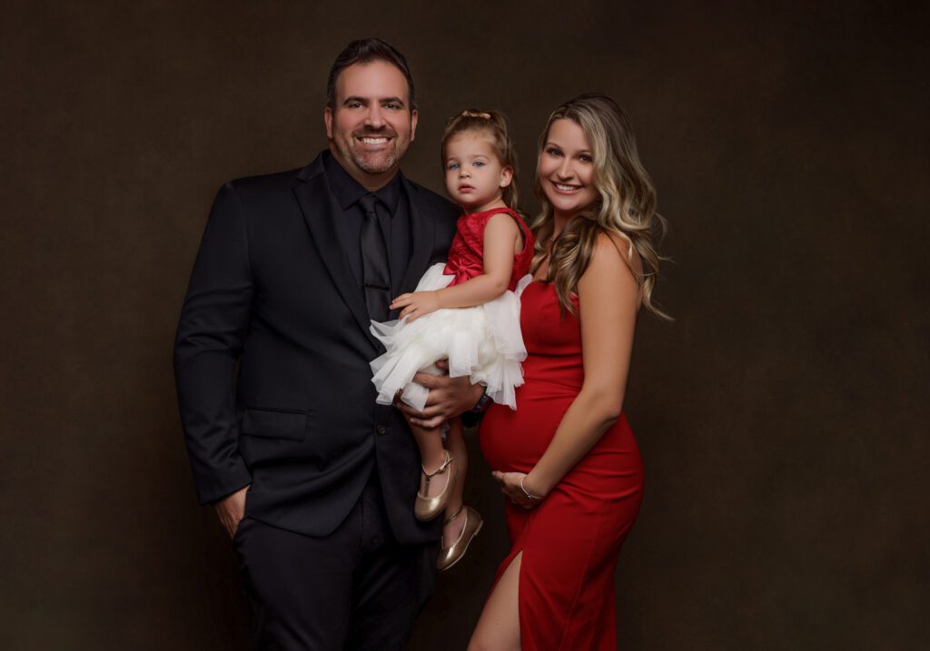 Pregnant woman standing with her partner and young child in a studio maternity portrait, photographed against a dark backdrop