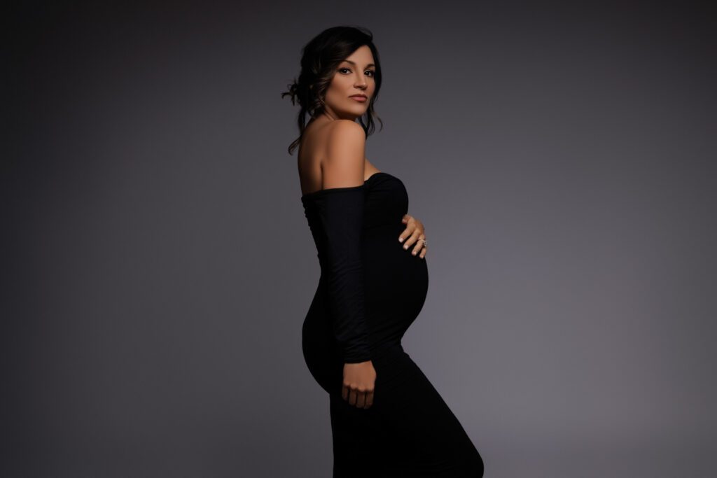 Pregnant woman in fitted black off-shoulder gown posing in studio maternity photography session in South Florida