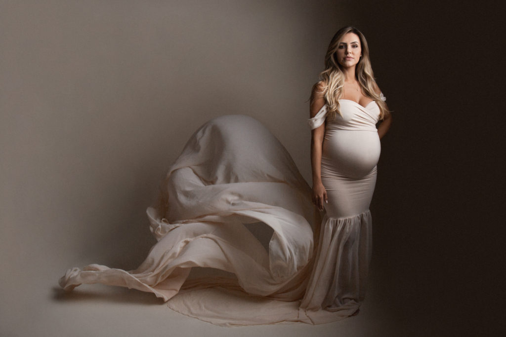 pregnant model wearing cream colored off-the-shoulder gown looks into camera lens as cream colored silk billows behind her