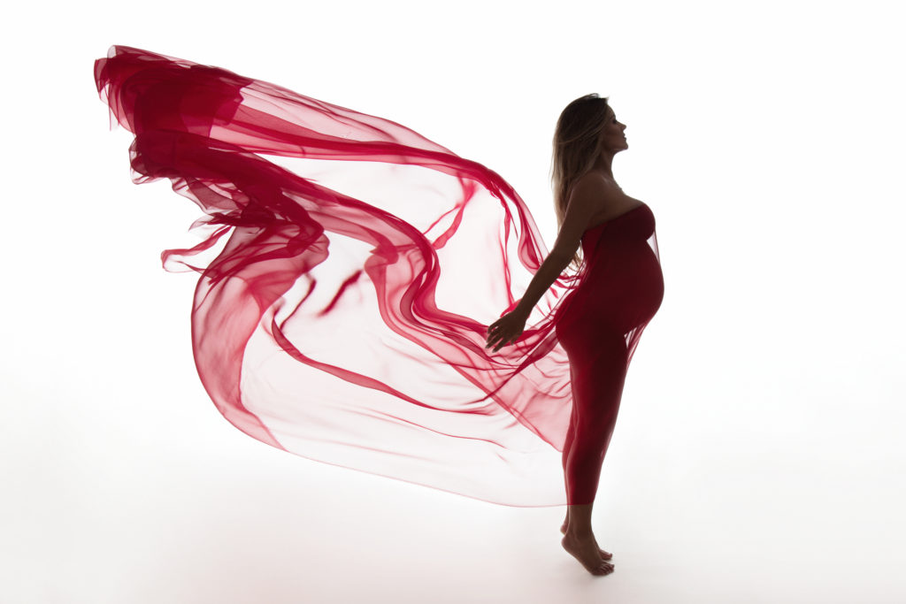 pregnant woman stands on tip toes with red silk billowing behind her