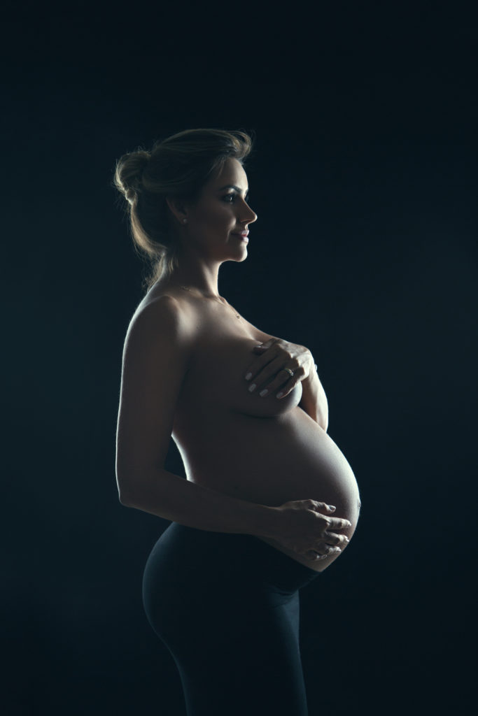 pregnant woman nude from the waste up covers her breasts with one hand and her belly with the other in soft profile color image