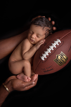newborn sleeping baby being held by two large, brown hands snuggles a football while in a fetal position