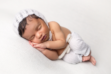 newborn baby sleeping on side with hands under his cheek, wearing a white knit cap and white bottoms against a white background