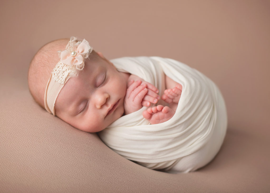 peaceful baby girl with an ivory headband swaddled in white cloth sleeping on a tan blanket