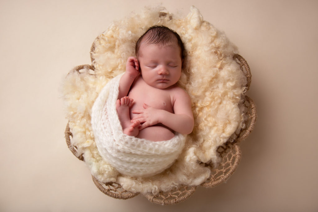 adorable baby boy in white fabric inside a basket on a tan background