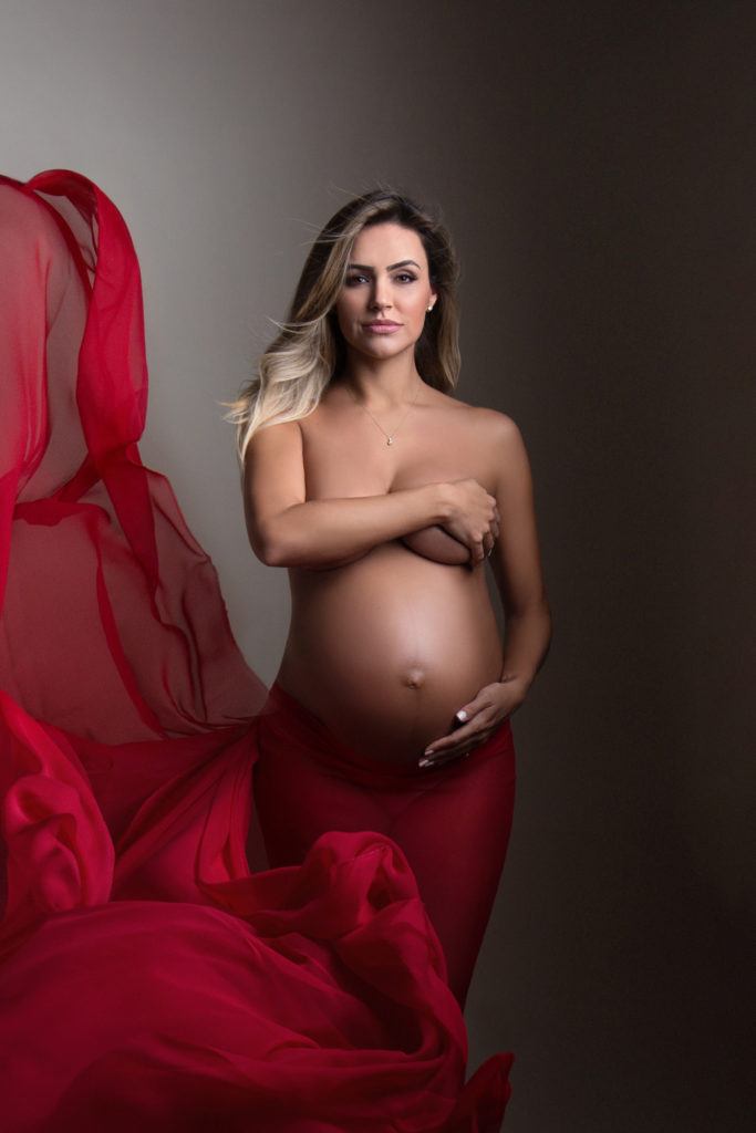 Pregnant mom in red blowing fabric smiles while holding her belly in maternity photography