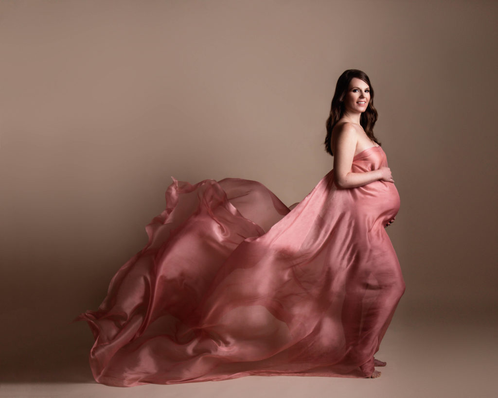 Pregnant woman in coral pink dress smiling and holding baby bump against beige background for maternity picture outfit ideas