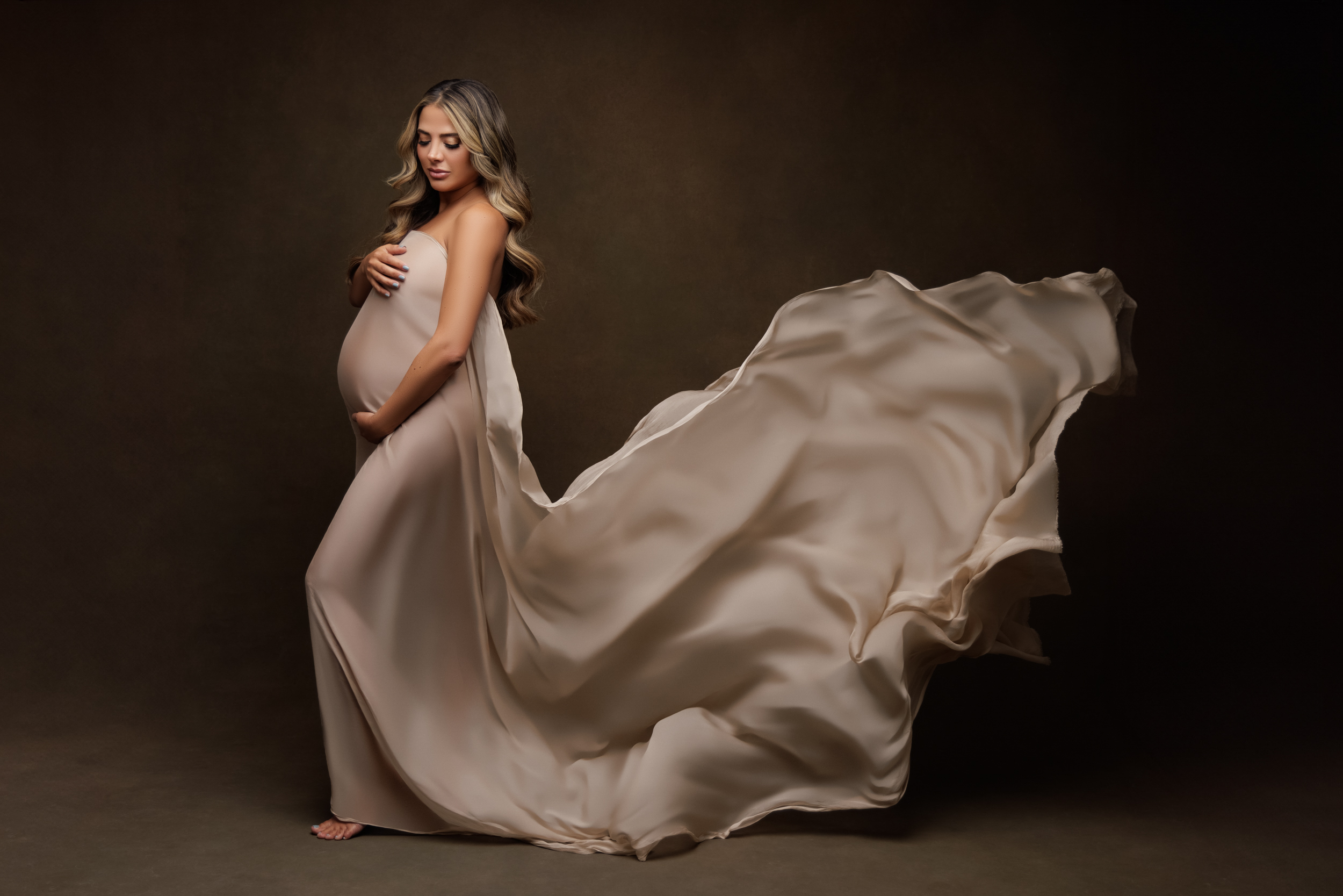 Pregnant woman standing in a studio wearing a flowing neutral gown, photographed in a maternity portrait with soft lighting