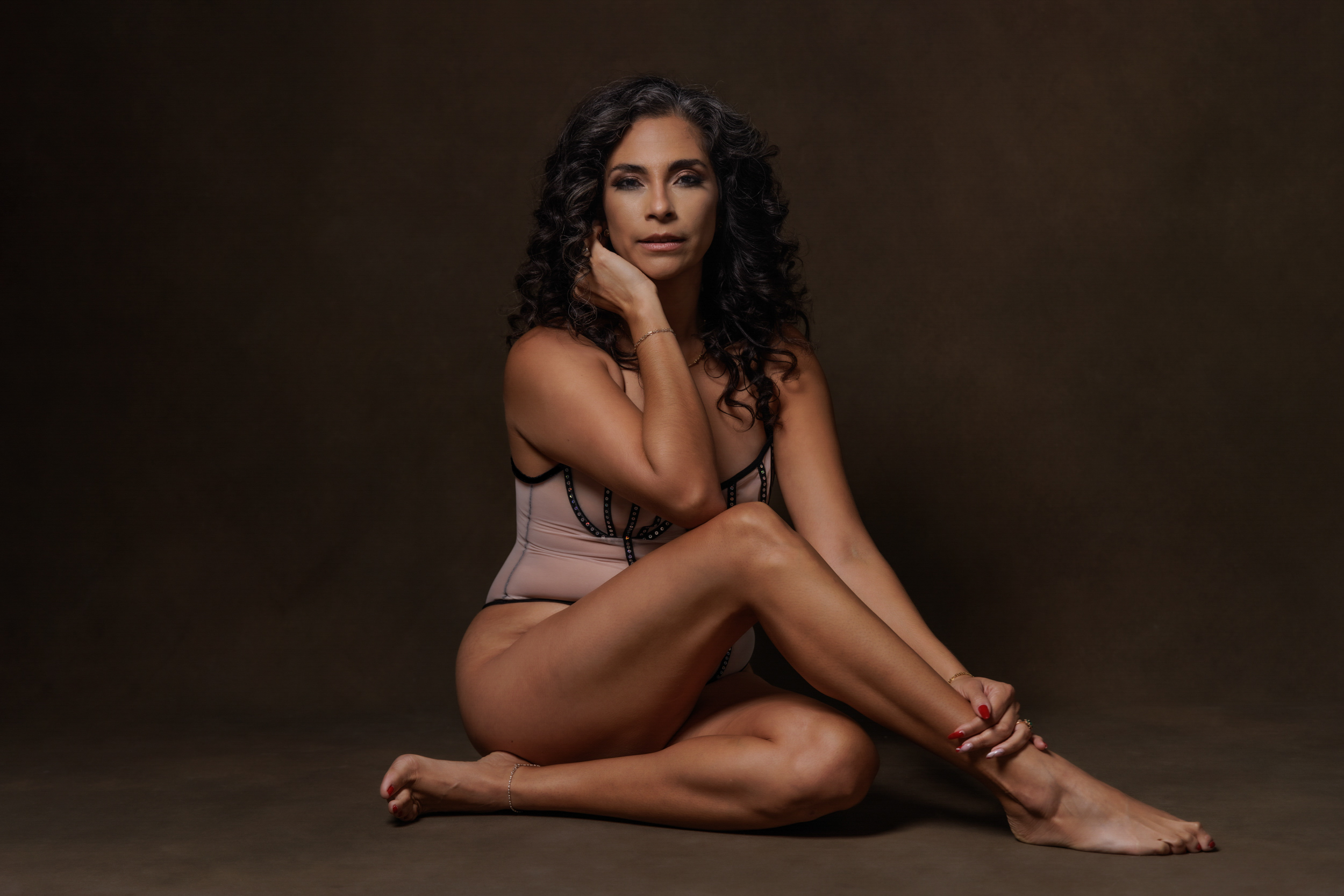 Woman seated on the floor wearing a blush bodysuit, photographed in a studio boudoir portrait with soft directional lighting in Miami Florida