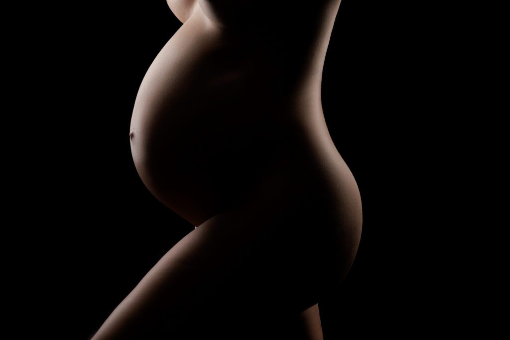Silhouette of pregnant woman in dramatic studio lighting showing sculpted belly and curves during fine art maternity photography session in Miami